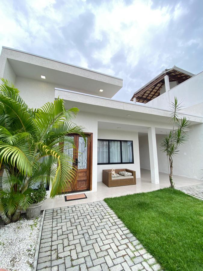 The exterior of the house features a welcoming entrance with a wooden door and a cozy seating area. Lush green plants and well-maintained grass frame the entry, while a stone pathway leads to the front door. The upper level contains an open terrace with a roof.