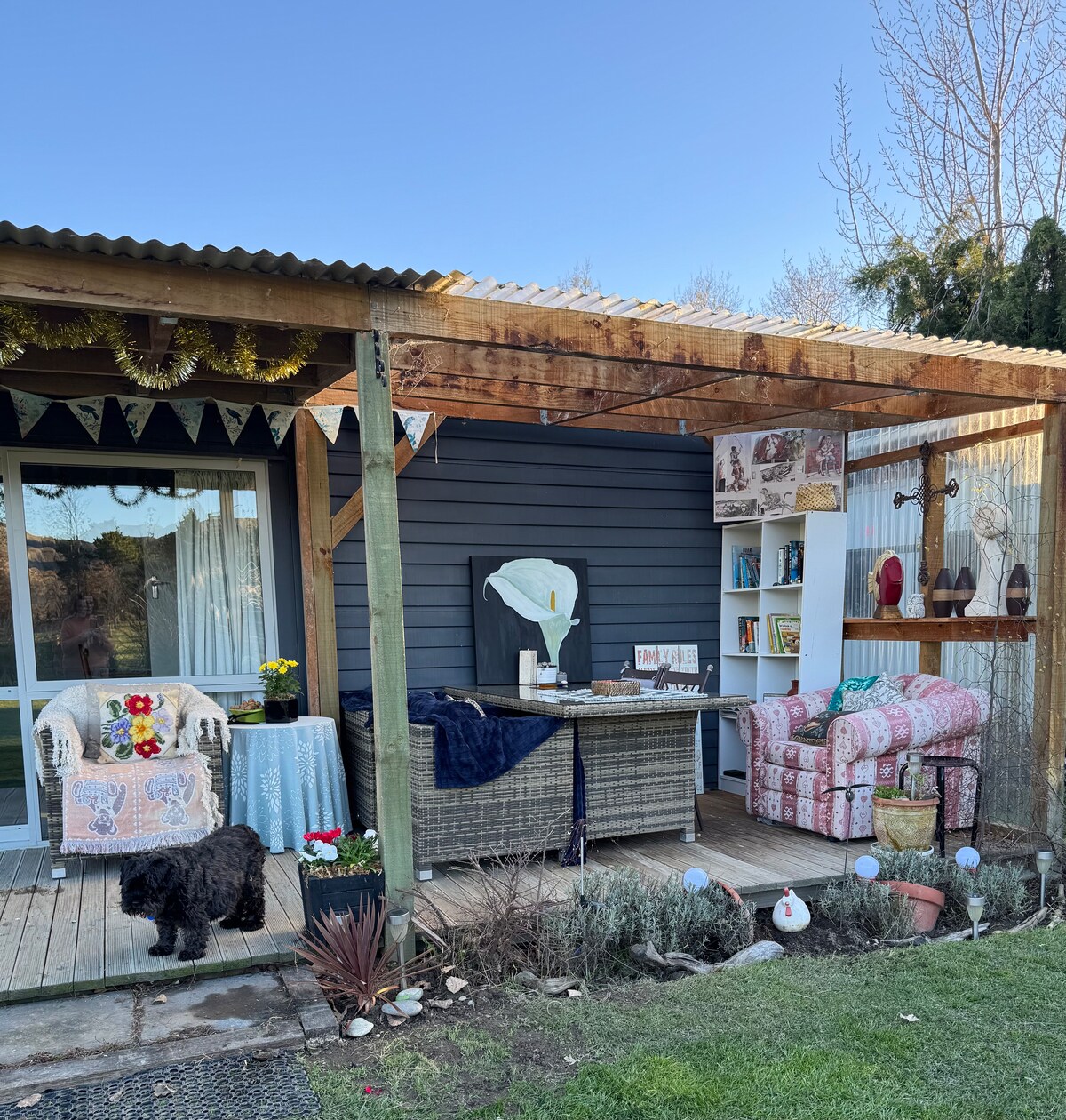 An outdoor seating area is shown with a cozy atmosphere, featuring a table adorned with a blue cloth. Two patterned chairs offer relaxation options. Plant beds line the edge, and a friendly black dog sits nearby. Decorative items and books are visible on a nearby shelf.