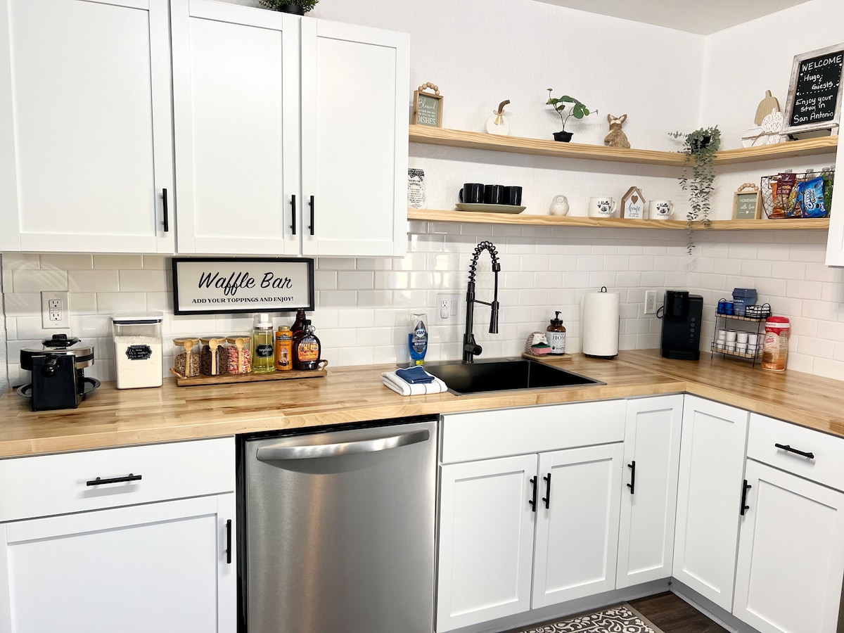 A bright kitchen features white cabinetry and wooden countertops, providing a clean and inviting space. A stainless steel sink is positioned next to a variety of kitchen essentials. Shelves display decorative items and organized containers, contributing to a well-equipped cooking environment.