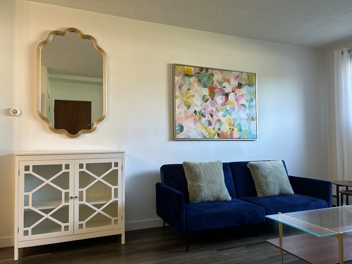 A stylish living space features a navy blue sofa adorned with two decorative pillows. A modern coffee table sits in front, and a light wood cabinet with geometric design is positioned nearby. An abstract painting in soft colors adds a touch of artistry on the wall.
