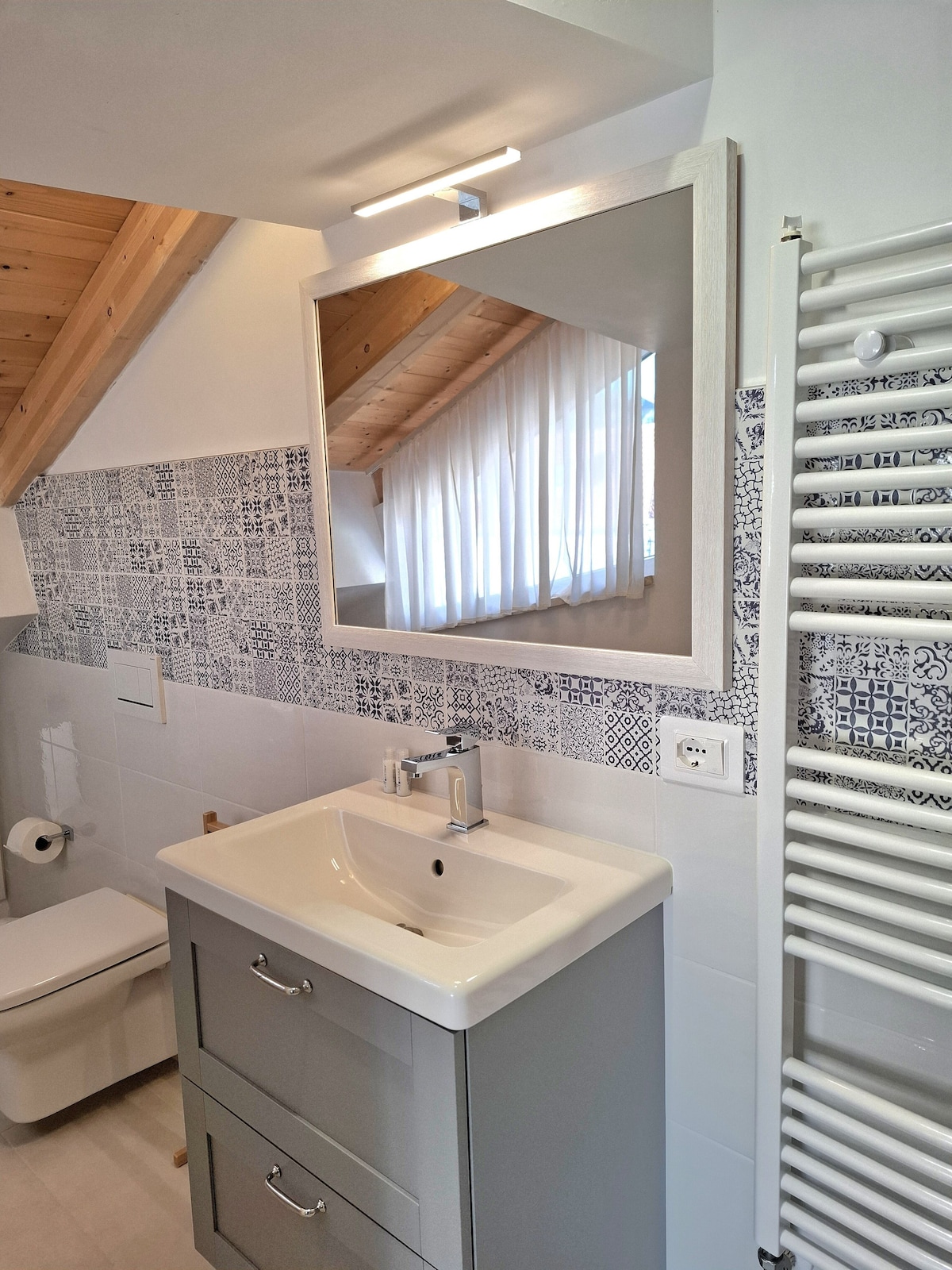 A modern bathroom features a sleek sink with a minimalist design set against light gray cabinetry. Above the sink, a large mirror is framed simply. Natural light filters through sheer curtains, illuminating the space. A towel radiator complements the aesthetic.