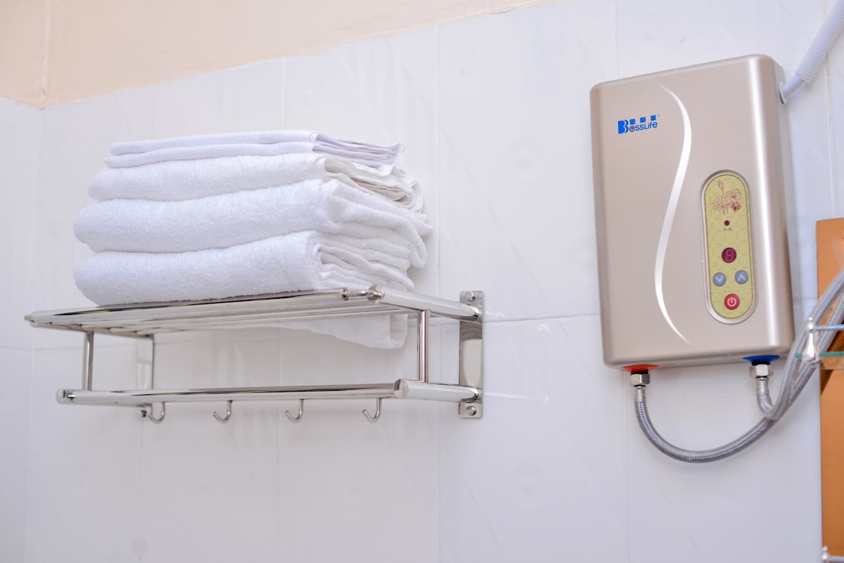 A clean and functional bathroom space is highlighted by neatly stacked white towels on a stainless steel shelf. A shower unit is mounted above, featuring a streamlined design with accessible controls for temperature adjustment.