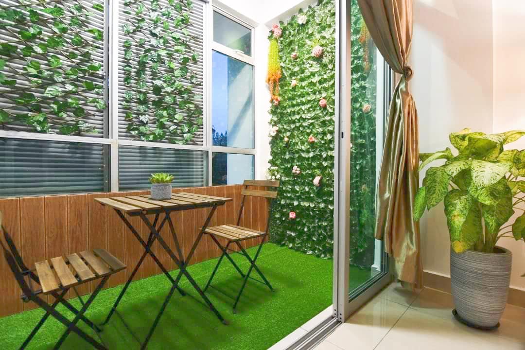 A cozy outdoor space features a small wooden table paired with two folding chairs on a green carpet. Lush greenery and floral decorations adorn the walls, creating a relaxing environment. A potted plant is placed in the corner, bringing a touch of nature indoors.