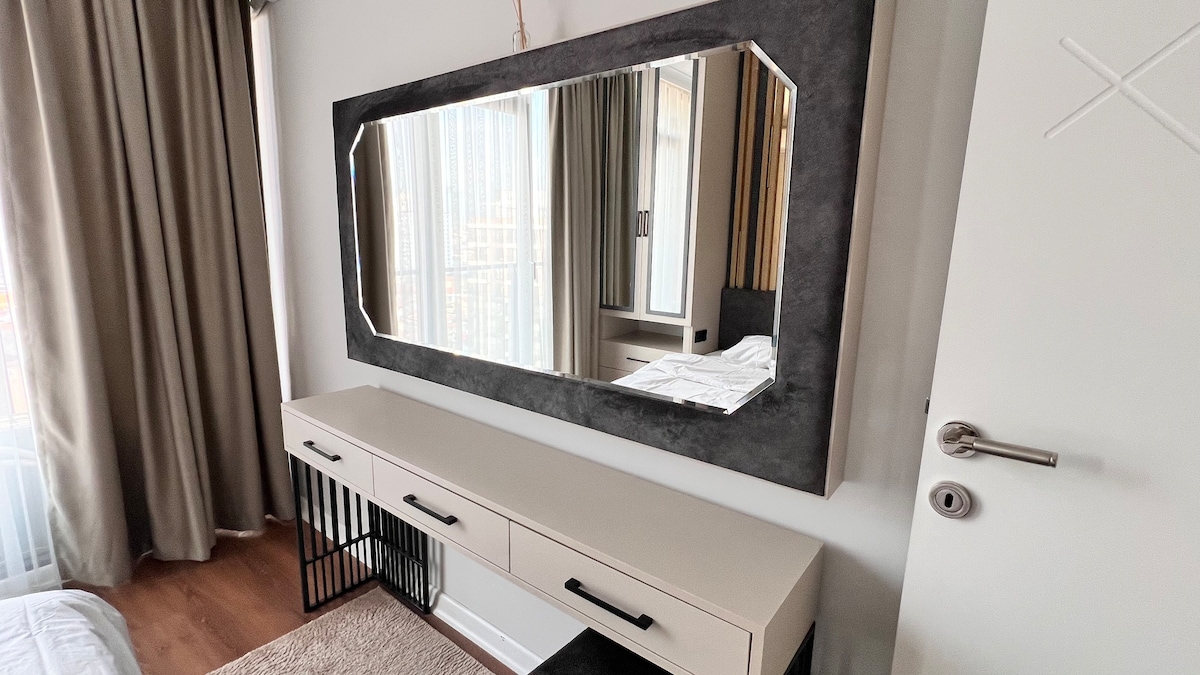 A sleek vanity table with three drawers is positioned against the wall. A large, rectangular mirror frames the table, reflecting light from a nearby window, accentuated by flowing curtains. The warm wood flooring contrasts with the modern furnishings, creating a harmonious aesthetic.