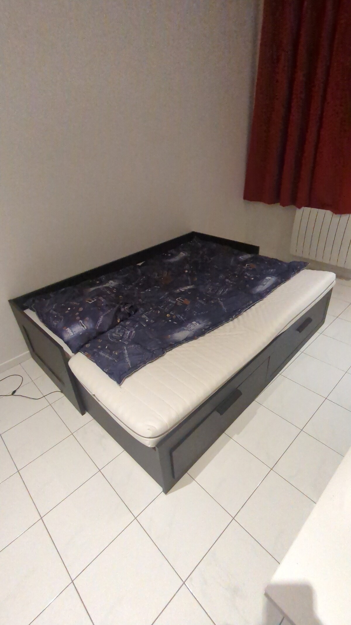 A compact bed frame is positioned against a plain wall, featuring a pull-out mattress covered with cozy bedding. The room includes a large window dressed with bold red curtains, contributing to a relaxed environment. A tiled floor adds a clean, modern feel.