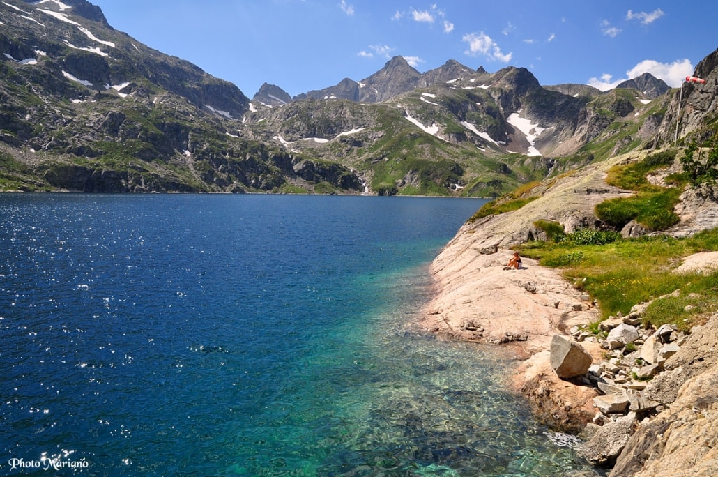 A serene alpine lake is bordered by rocky shores and lush greenery. The clear water transitions from deep blue to vibrant turquoise near the shoreline, reflecting the surrounding mountains topped with patches of snow. A few scattered rocks and plants are visible along the water's edge.