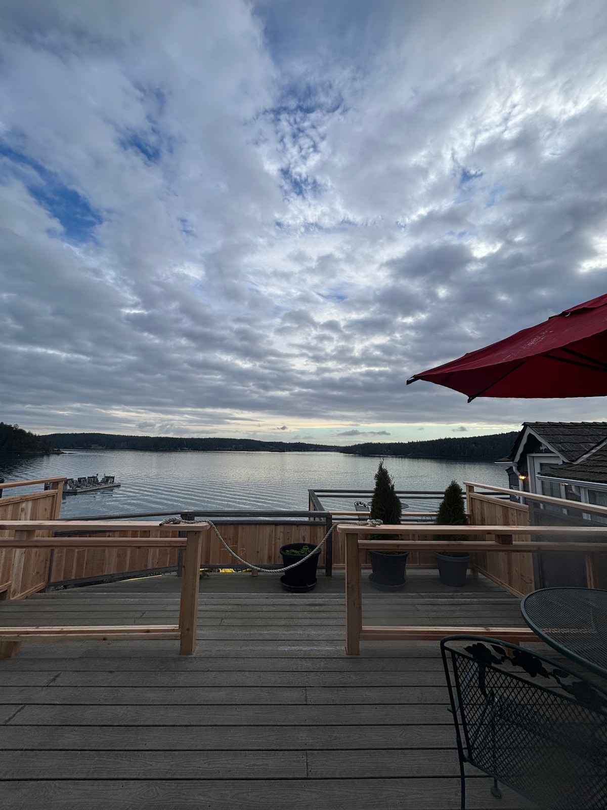 A spacious wooden deck extends over tranquil waters, framed by lush, evergreen trees. A large umbrella provides shade, while a small bistro table and chairs invite relaxation. The sky is filled with soft, gray clouds, reflecting the calm ambience of the waterfront setting.