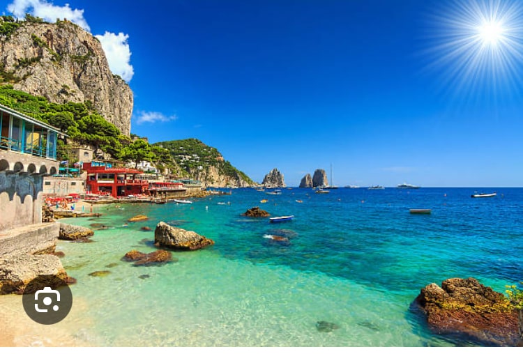 A coastal view displays a vibrant turquoise sea lapping against rocky shores. Cliffs rise in the background, dotted with greenery, while boats float in the clear water. Bright sun illuminates the scene, highlighting the inviting atmosphere of this popular seaside destination.