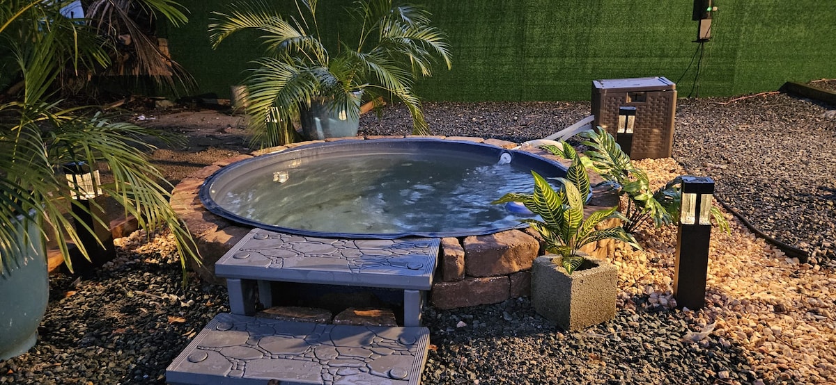 A circular hot tub is nestled among greenery, surrounded by decorative plants in pots. Steps lead up to the tub, which is illuminated by soft lighting from lanterns nearby. The space features pebbled ground, providing a natural and relaxed environment.