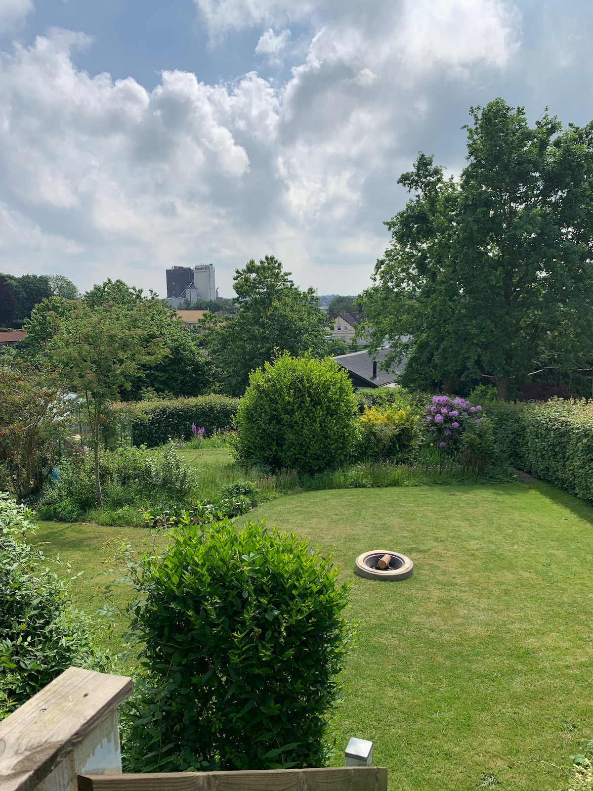 A lush garden is surrounded by various trees and shrubs, providing a sense of privacy. A circular seating area is visible on the well-maintained grass, with flowering plants adding color. The sky is partly cloudy, allowing soft light to filter through the greenery.