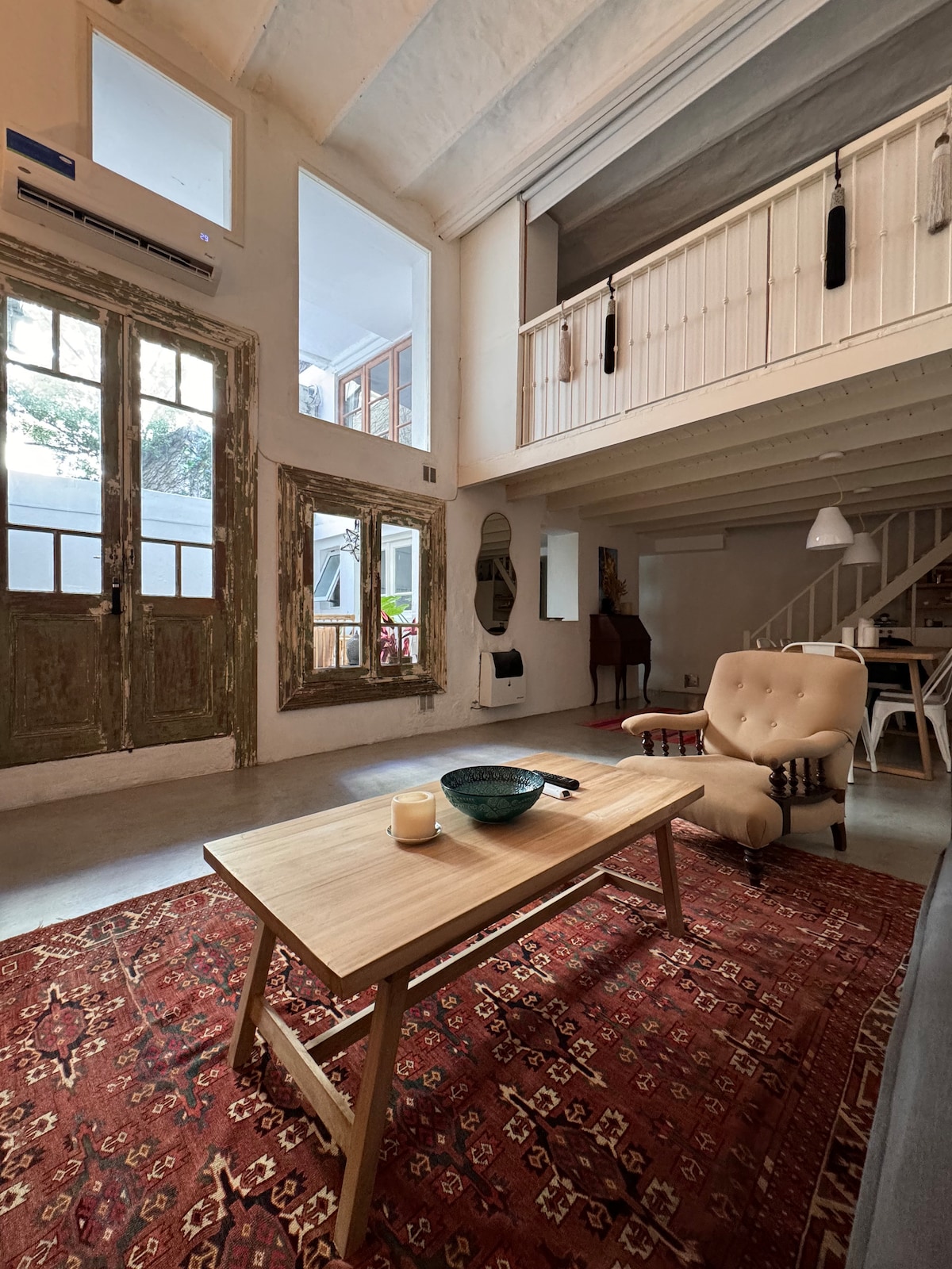 The spacious living area features a high ceiling with large windows allowing natural light. A wooden table sits at the center, complemented by an armchair and a decorative rug. The space connects to a patio visible through the glass doors.
