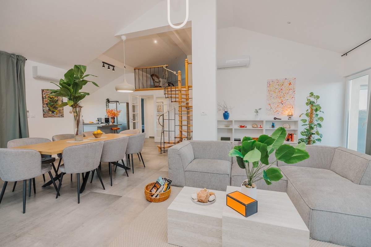 A spacious interior features modern furnishings and an open-concept layout. A comfortable sectional sofa is positioned around a light-colored coffee table, with a dining area nearby showcasing a round table and upholstered chairs. Natural light streams in through large windows, highlighting decorative plants.