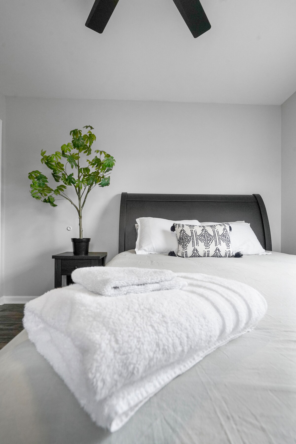 A well-lit bedroom features a spacious bed made with soft linens and decorative pillows. A small potted plant adds a touch of greenery. A neatly folded set of towels rests on the bed, complemented by a ceiling fan above.