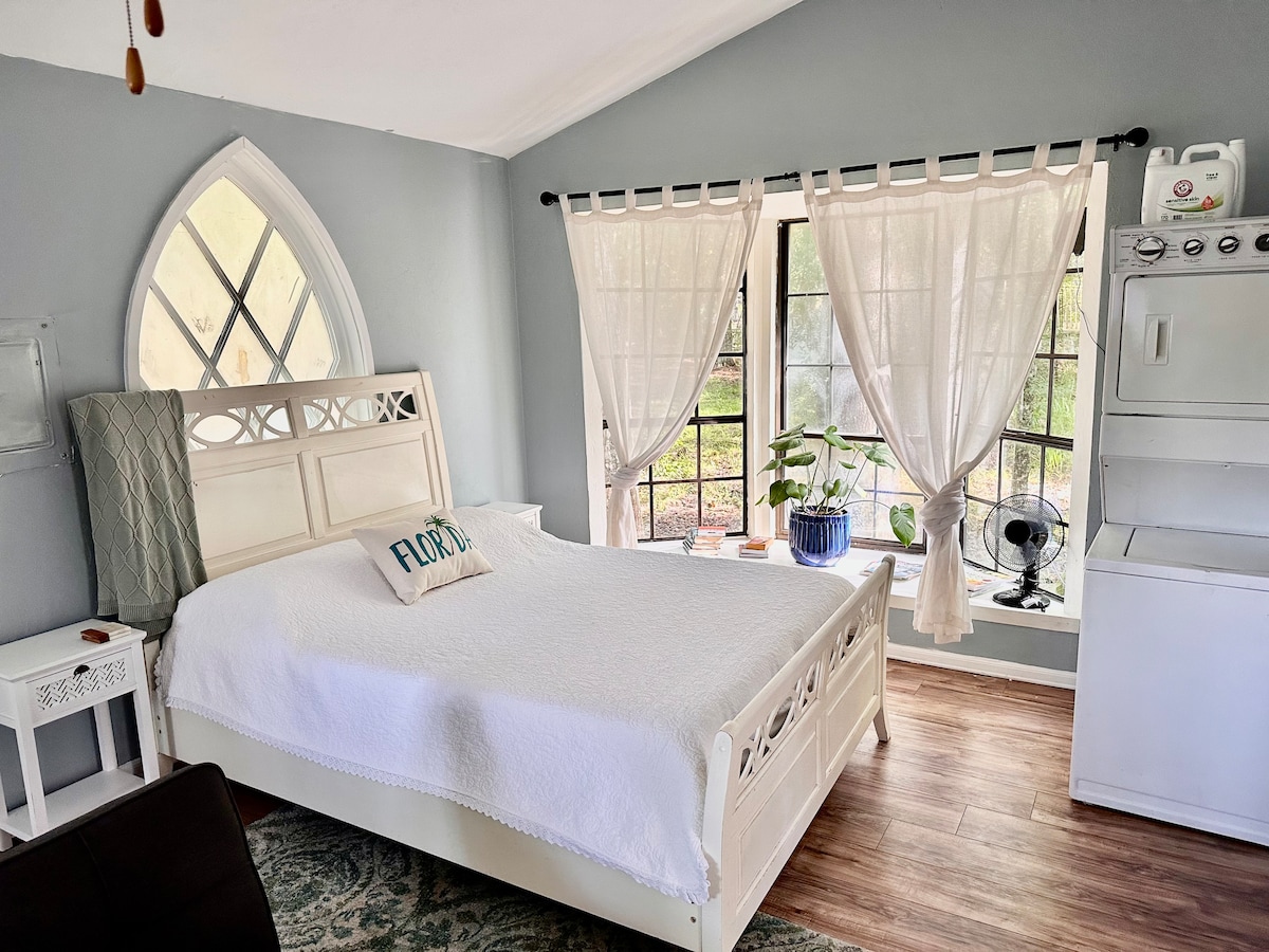 A studio space features a queen-sized bed with a light blanket and decorative pillow. Large windows allow natural light to fill the room, partially obscured by sheer curtains. A small fan and potted plant enhance the cozy setting, complemented by a compact kitchen area.