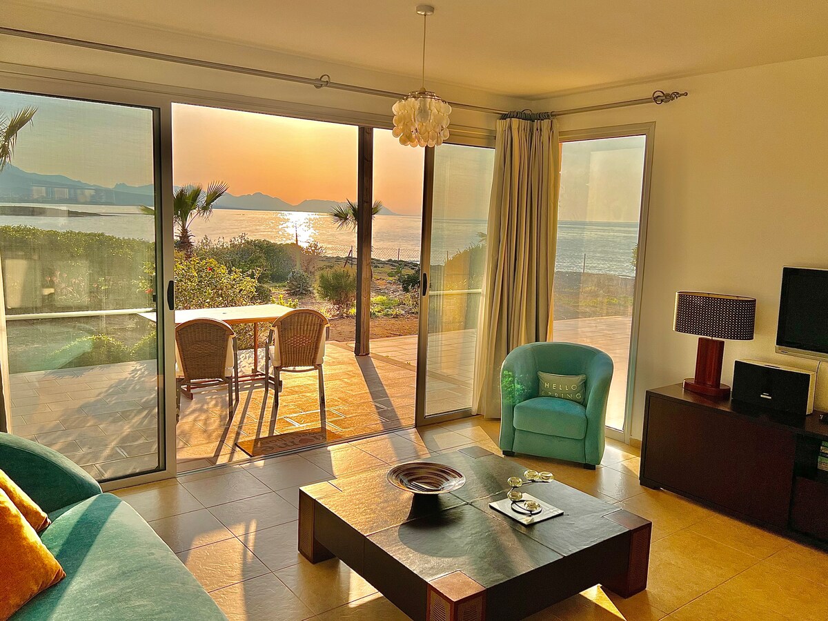 A living area is bathed in warm sunset light, featuring large glass doors that open to a serene outdoor view. A table with two chairs is positioned on the terrace outside, while a soft blue-green chair adds a pop of color to the neutral decor.