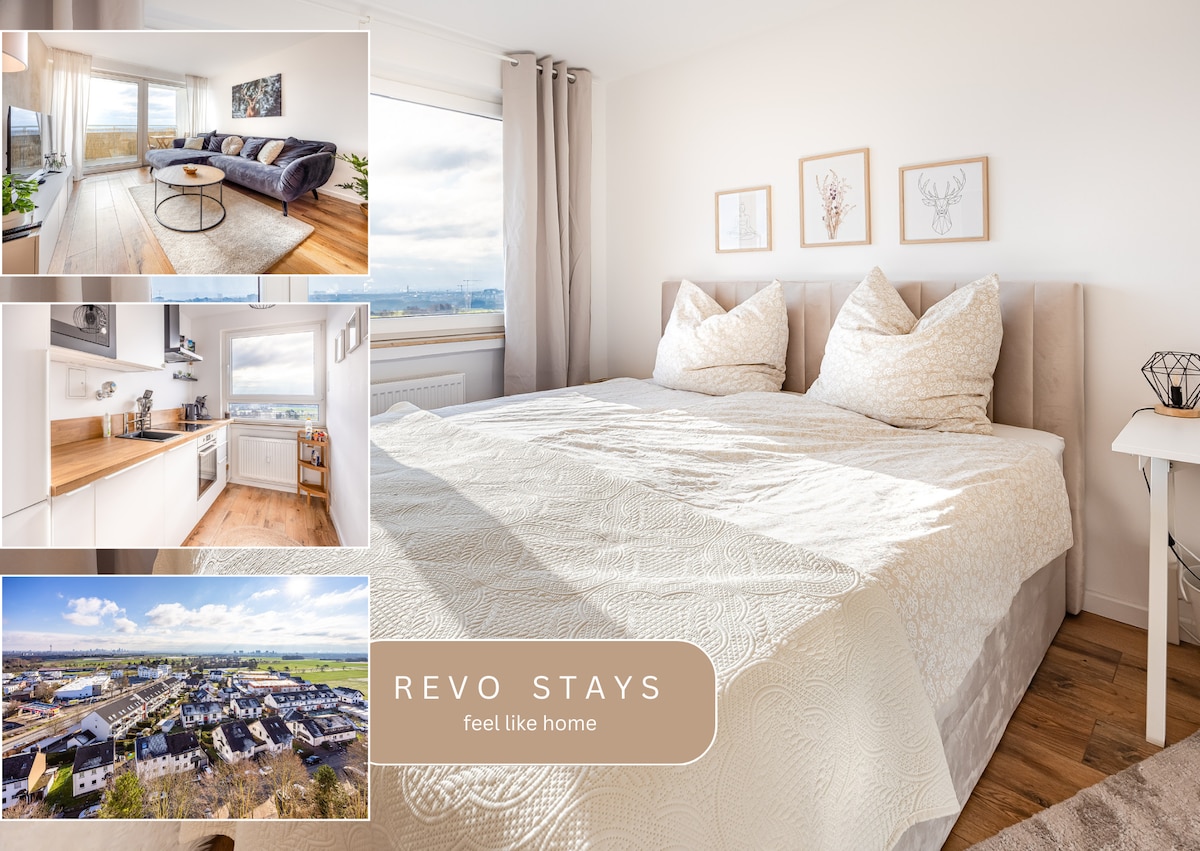 A well-lit bedroom features a spacious bed with decorative pillows and a light-colored coverlet. Framed artwork adorns the wall above the bed, while large windows provide a panoramic view of the surroundings, allowing ample natural light to fill the space.