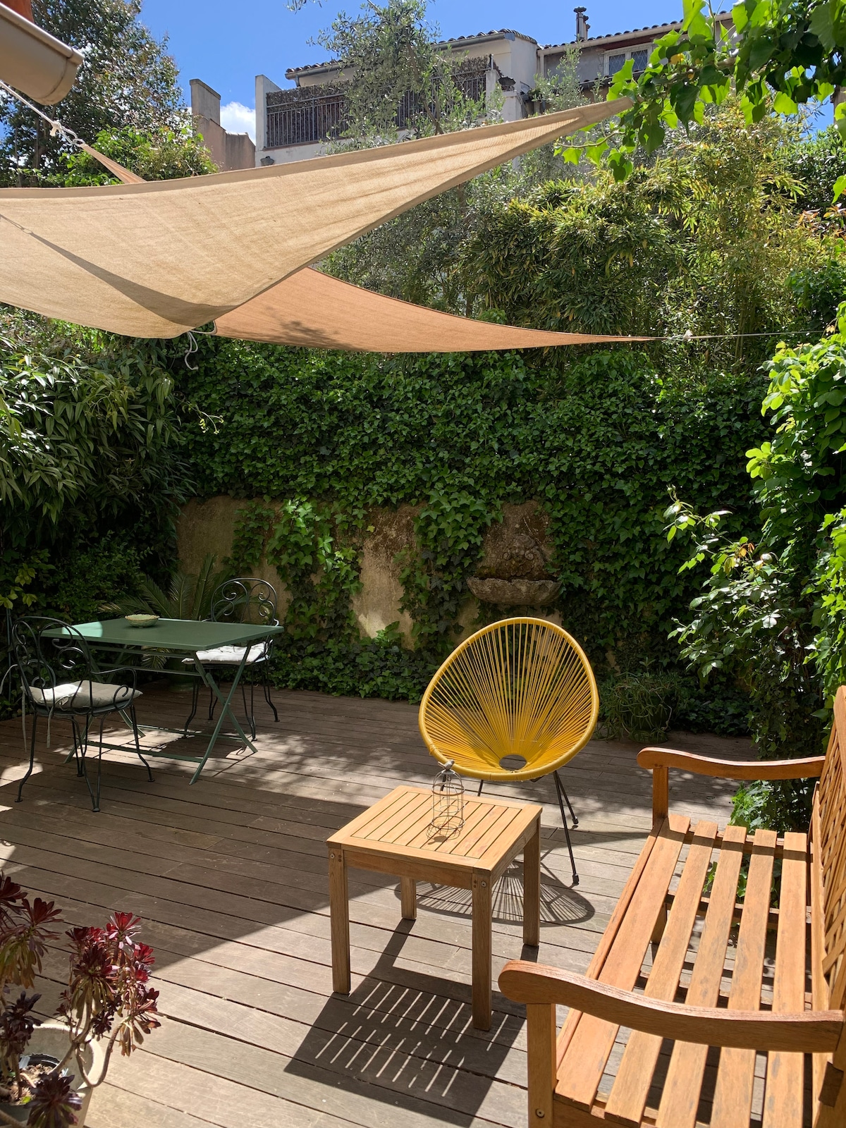 A private garden area features wooden decking surrounded by greenery. A yellow chair and wooden bench are situated beside a small table. A metal dining table is also visible, shaded by fabric canopies that provide a relaxed outdoor space.
