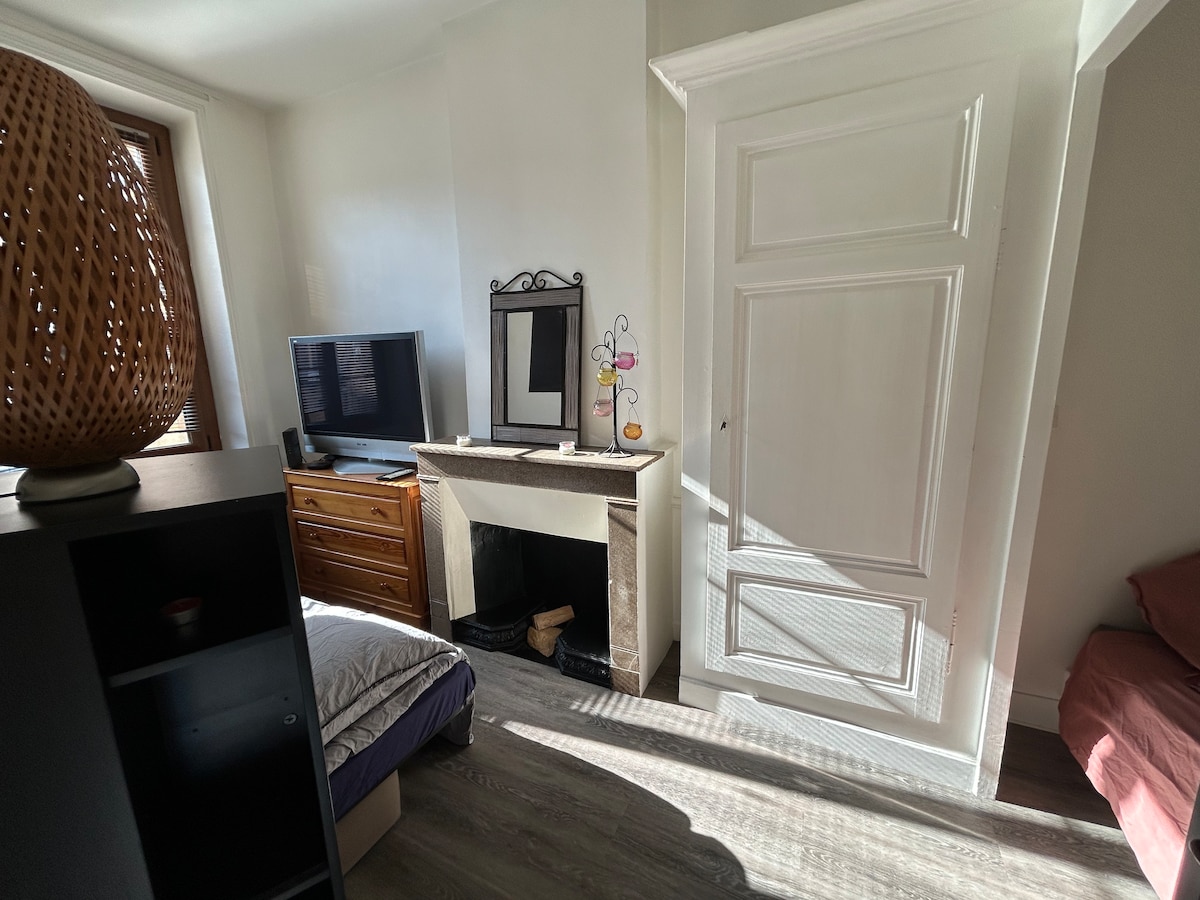 A cozy corner of the apartment features a light-filled space with a wooden dresser and a flat-screen television. A decorative fireplace is situated beneath a mirrored frame, while a spacious bed with a neatly arranged bedspread is positioned nearby.