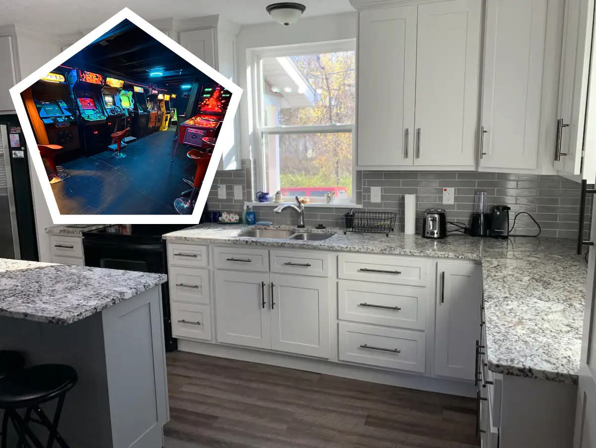 A modern kitchen features a spacious granite countertop and light cabinetry. Natural light from a nearby window brightens the space. An inset view displays a vibrant arcade area with several vintage arcade games, creating a playful contrast to the kitchen's sleek design.
