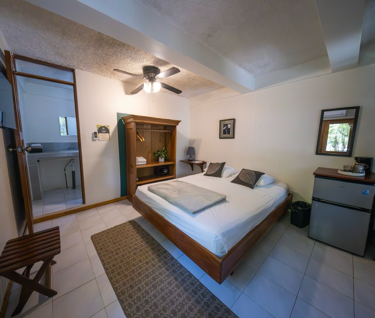 A spacious sleeping area features a large bed with neutral bedding and accent pillows. A fan hangs from the ceiling, providing comfort. To one side, a mini refrigerator and small coffee station are present. A window allows natural light to enter, enhancing the calming atmosphere.