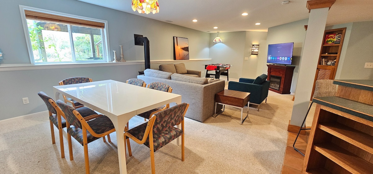 A spacious living area features a bright dining table with chairs, a cozy gray sofa, and a media unit with a large television. Soft lighting creates a relaxed environment, while a window allows natural light to illuminate the space. The carpeted floor adds warmth to the modern decor.