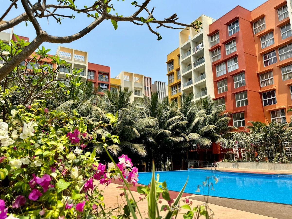 A clear blue swimming pool is framed by vibrant tropical gardens filled with blooming flowers. Colorful apartment buildings rise in the background, showcasing a mixture of red and yellow facades. Lush palm trees provide shade, creating a serene outdoor atmosphere.