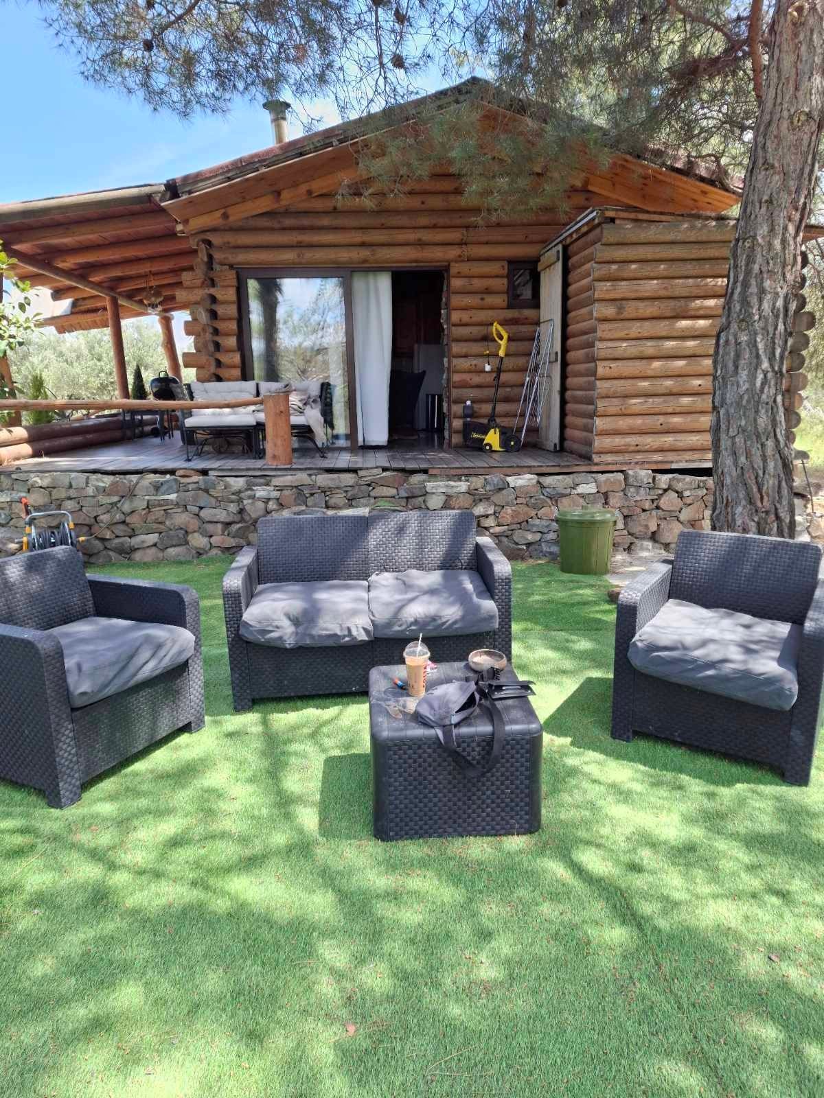 A comfortable outdoor seating area is arranged with four stylish wicker chairs surrounding a small table. The rustic wooden structure of the cabin is visible in the background, framed by lush greenery and natural stone elements.