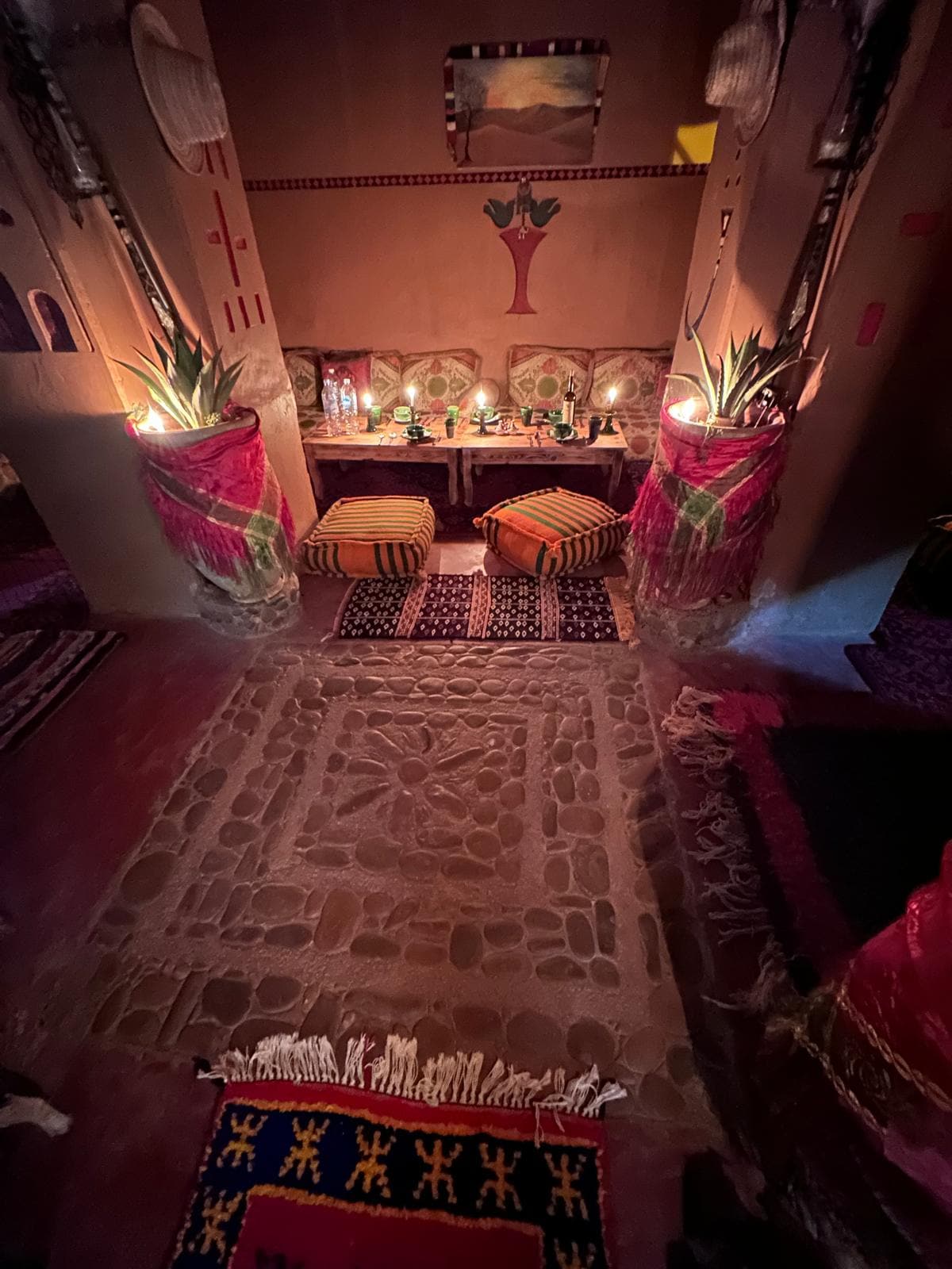A cozy sitting area features colorful cushions and traditional Moroccan textiles arranged around a low table. The space is softly lit by candles, creating a warm ambiance. The floor is decorated with patterned rugs, while decorative plants accentuate the inviting setting.