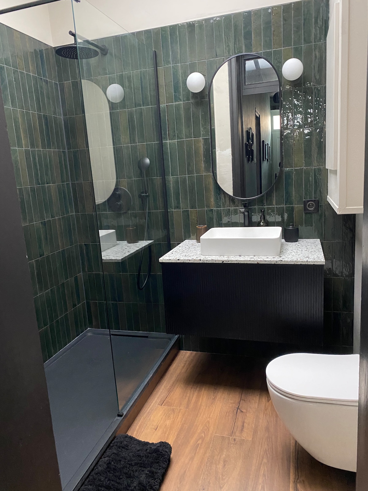 A modern bathroom features a spacious shower area enclosed by glass. The green tiled walls complement the minimalist vanity, topped with a square sink. A sleek toilet is positioned nearby, while warm wood flooring adds a touch of comfort to the space.