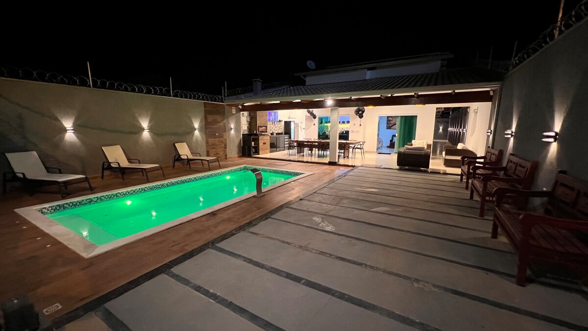 The outdoor area features a spacious pool with green illumination, surrounded by deck chairs. A large, covered area showcases a dining space and kitchen, with seating arrangements made of wooden benches. Soft lighting enhances the ambiance during the evening.
