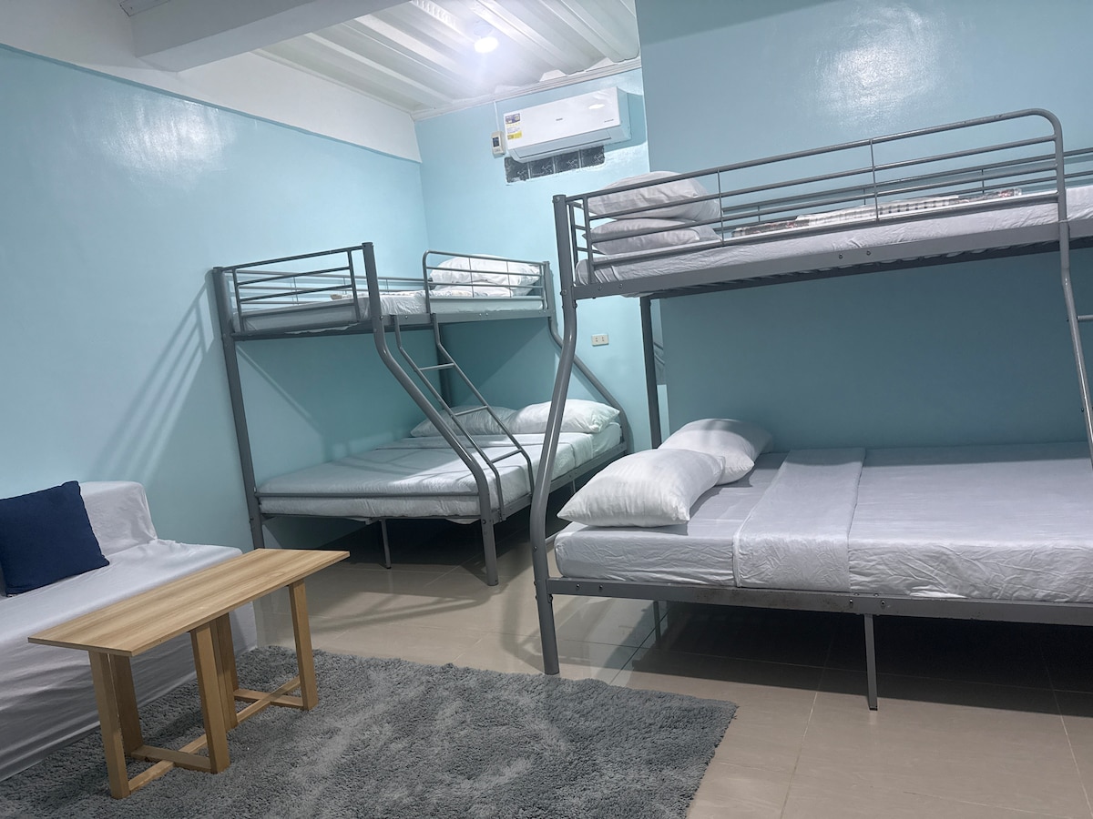 The sleeping area is furnished with two sets of metal bunk beds alongside a spacious bed. A cozy seating area features a small wooden table, and a soft rug covers the floor, enhancing comfort. Light blue walls contribute to a bright and airy ambiance.