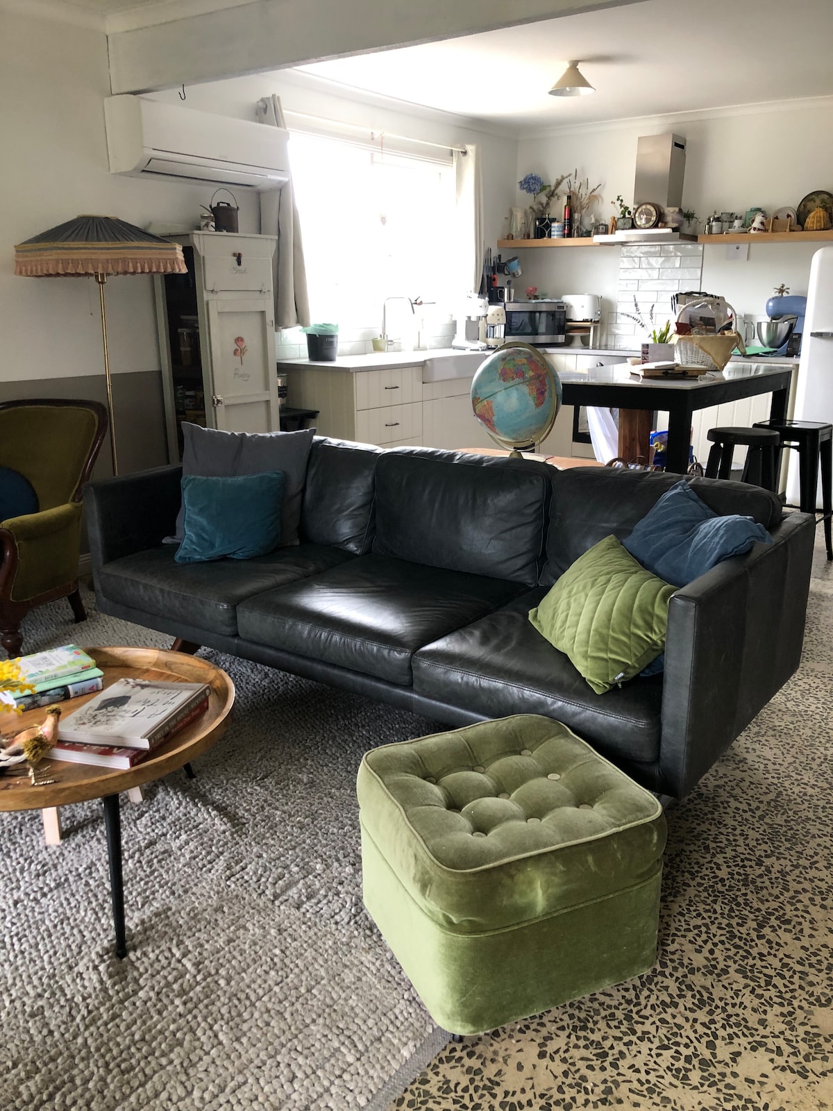 A comfortable living area features a dark leather sectional sofa adorned with various cushions. A round wooden coffee table holds magazines and flowers. A plush green ottoman complements the space, while a kitchen is visible in the background, showcasing a bright and functional design.