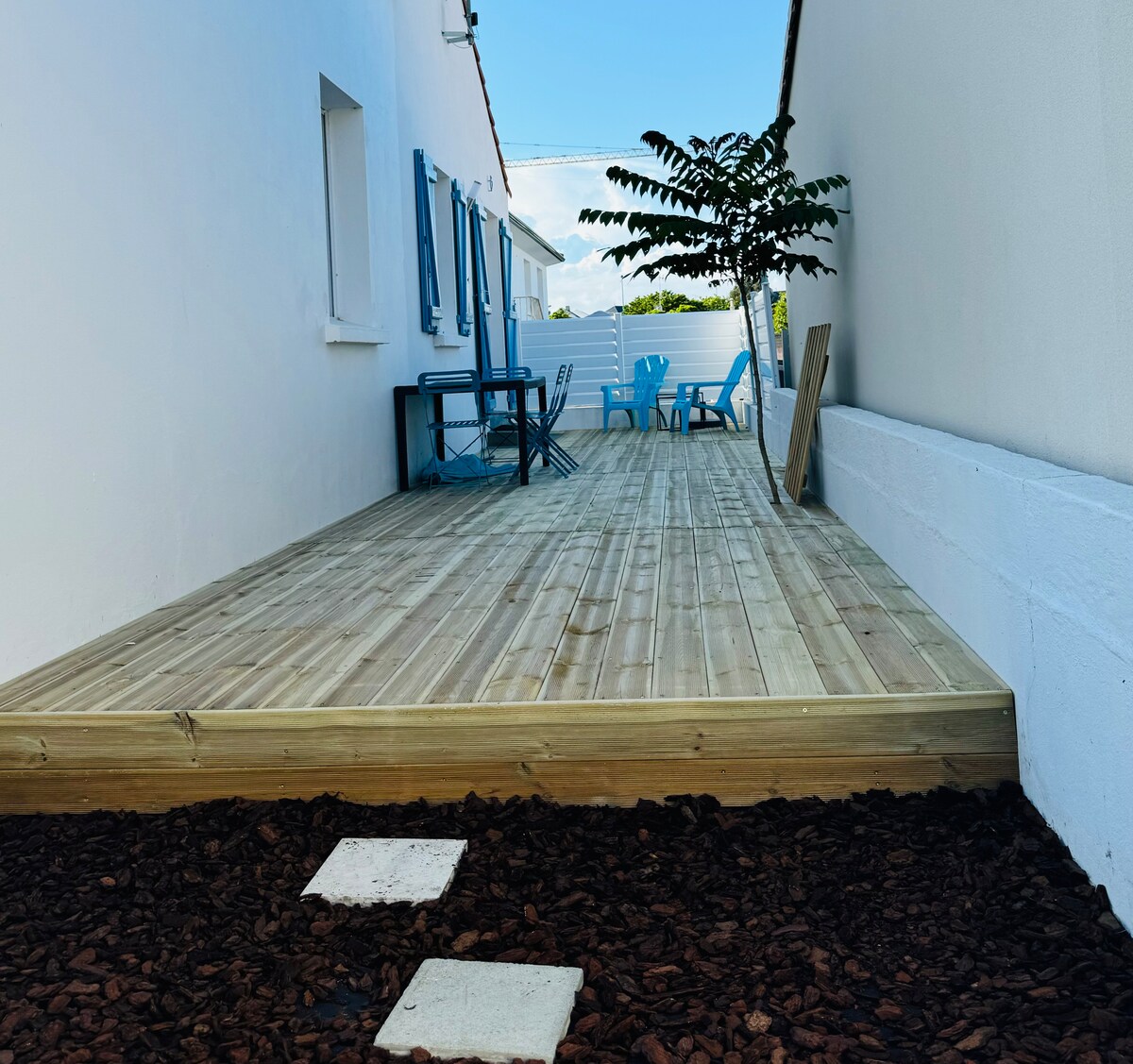 A spacious wooden terrace is presented, featuring a variety of outdoor seating options. Blue chairs and a small table offer a place to relax. The area is bordered by smooth stones and a lush green plant adds a touch of nature. The exterior walls are white.