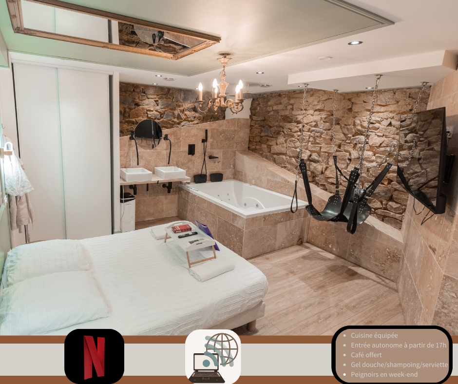 A cozy studio features a spacious bed, positioned near a unique hydro massage bathtub. Exposed stone walls add character, while soft lighting creates a relaxing ambiance. Two bathroom sinks are visible, along with specialized seating designed for comfort. A television is mounted on the wall for entertainment.
