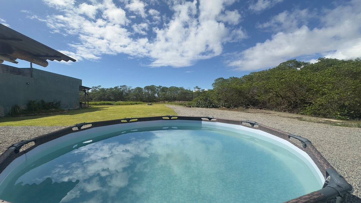 A round pool is shown in the foreground, reflecting the blue sky and scattered clouds. The background features a spacious green lawn bordered by trees, enhancing the natural setting of the property.