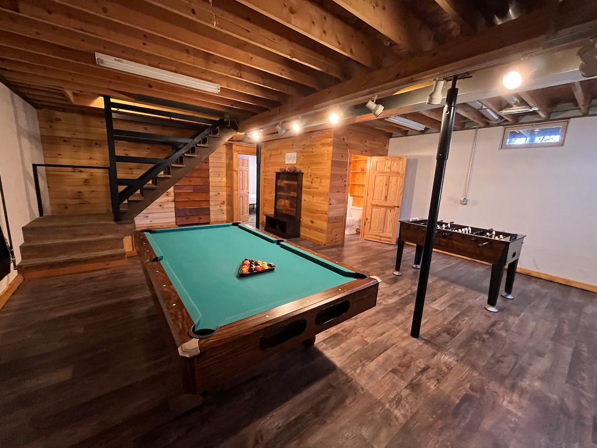 A game room features a green felt pool table with a set of billiard balls arranged on its surface. A foosball table stands in the corner, and wooden beams highlight the ceiling, creating a rustic feel. Natural light enters through a small window.