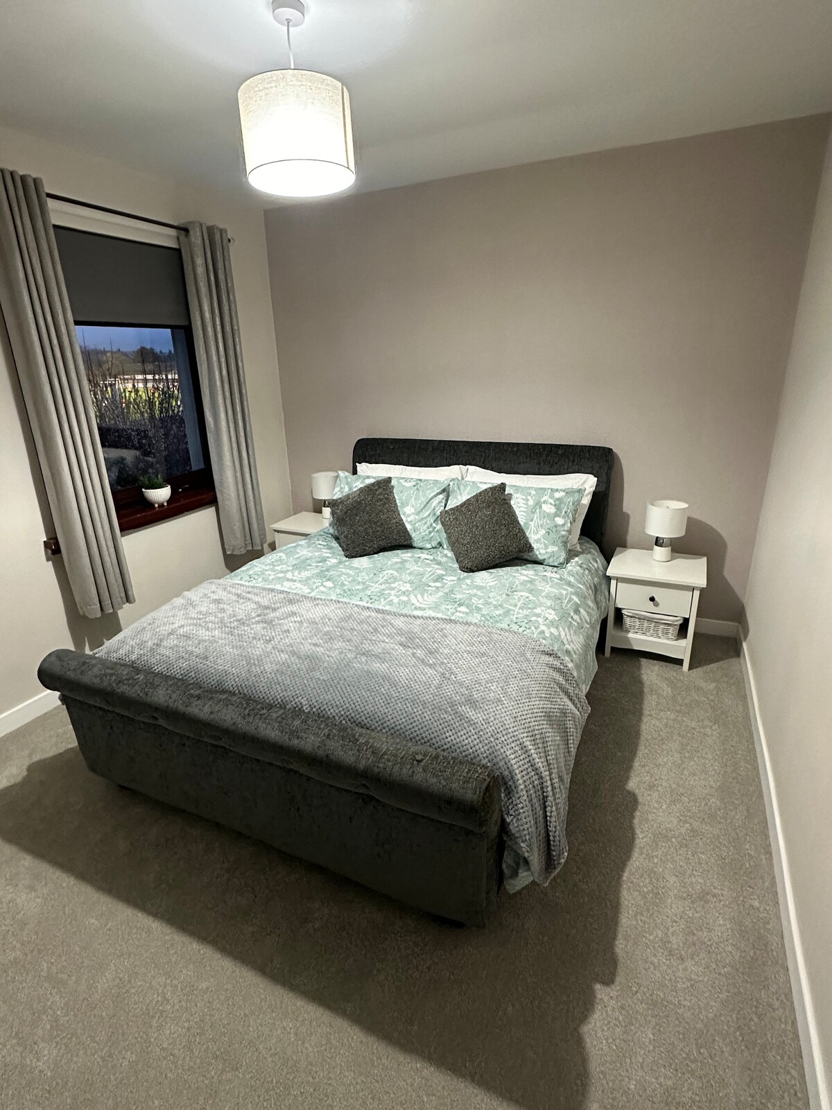 A well-appointed bedroom features a spacious bed dressed in a soft teal coverlet and coordinating pillows. Two bedside tables with lamps are positioned on either side. Natural light enters through a window, complemented by soft, light-grey curtains.