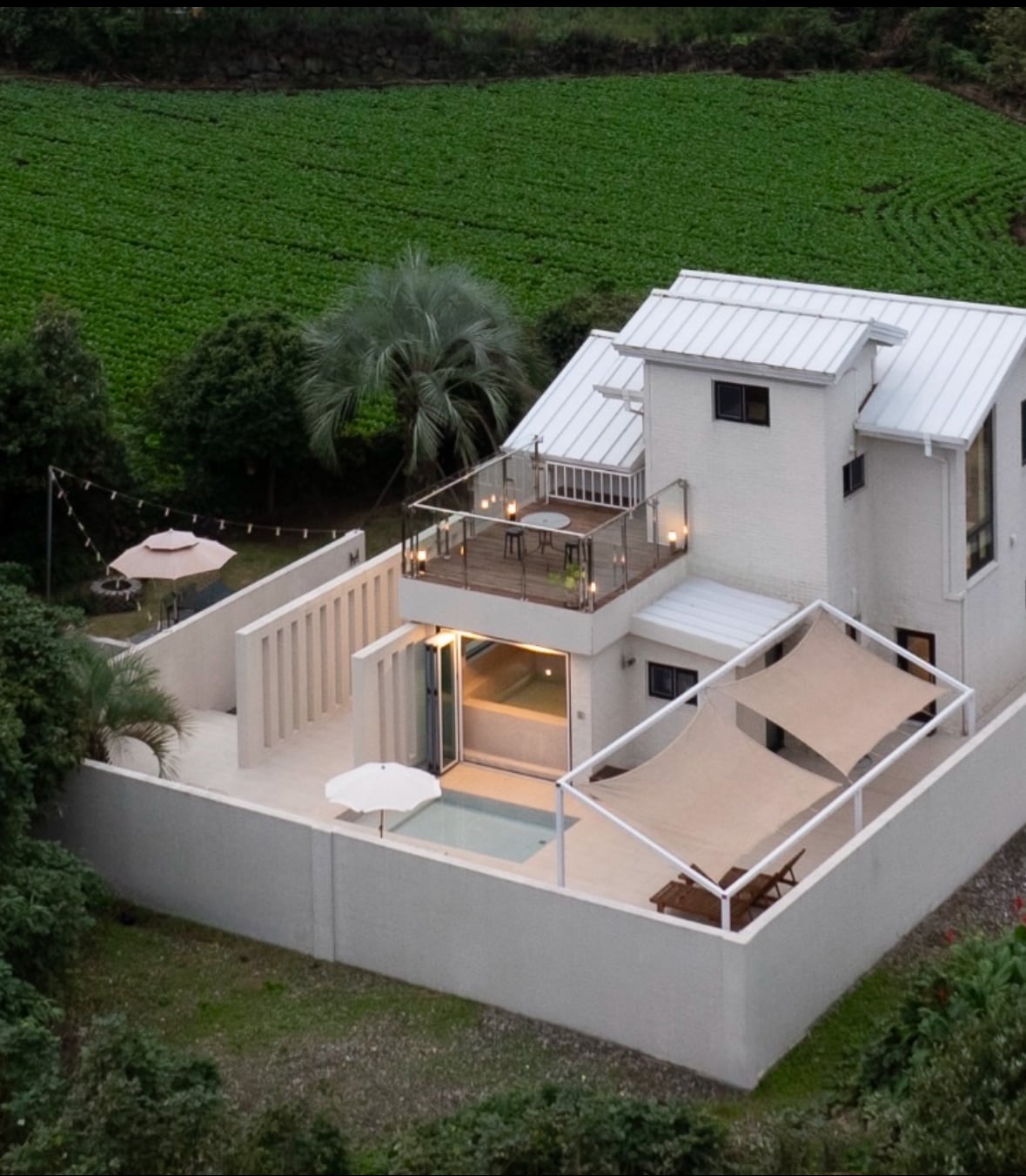 An aerial view of the property is displayed, showcasing a modern building surrounded by lush greenery. A spacious outdoor terrace with seating is visible, along with an umbrella and a shaded area. The pool area is highlighted, emphasizing the tranquil ambiance of the surroundings.
