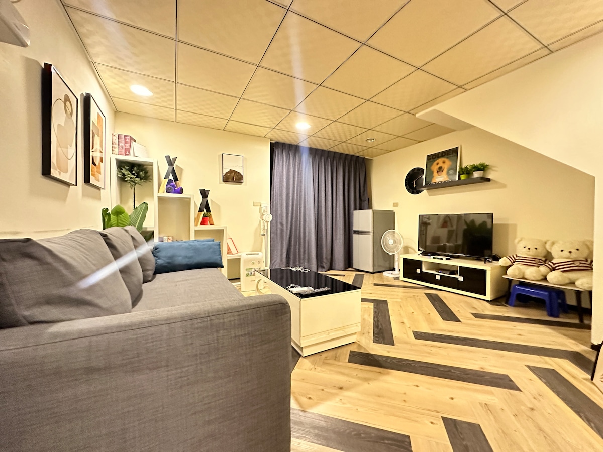 A cozy lounge area features a comfortable gray sofa and a minimalist coffee table, complemented by decorative shelves. A large flat-screen TV is positioned against the wall, and artwork is displayed nearby. Natural light filters in through the curtains, enhancing the inviting space.