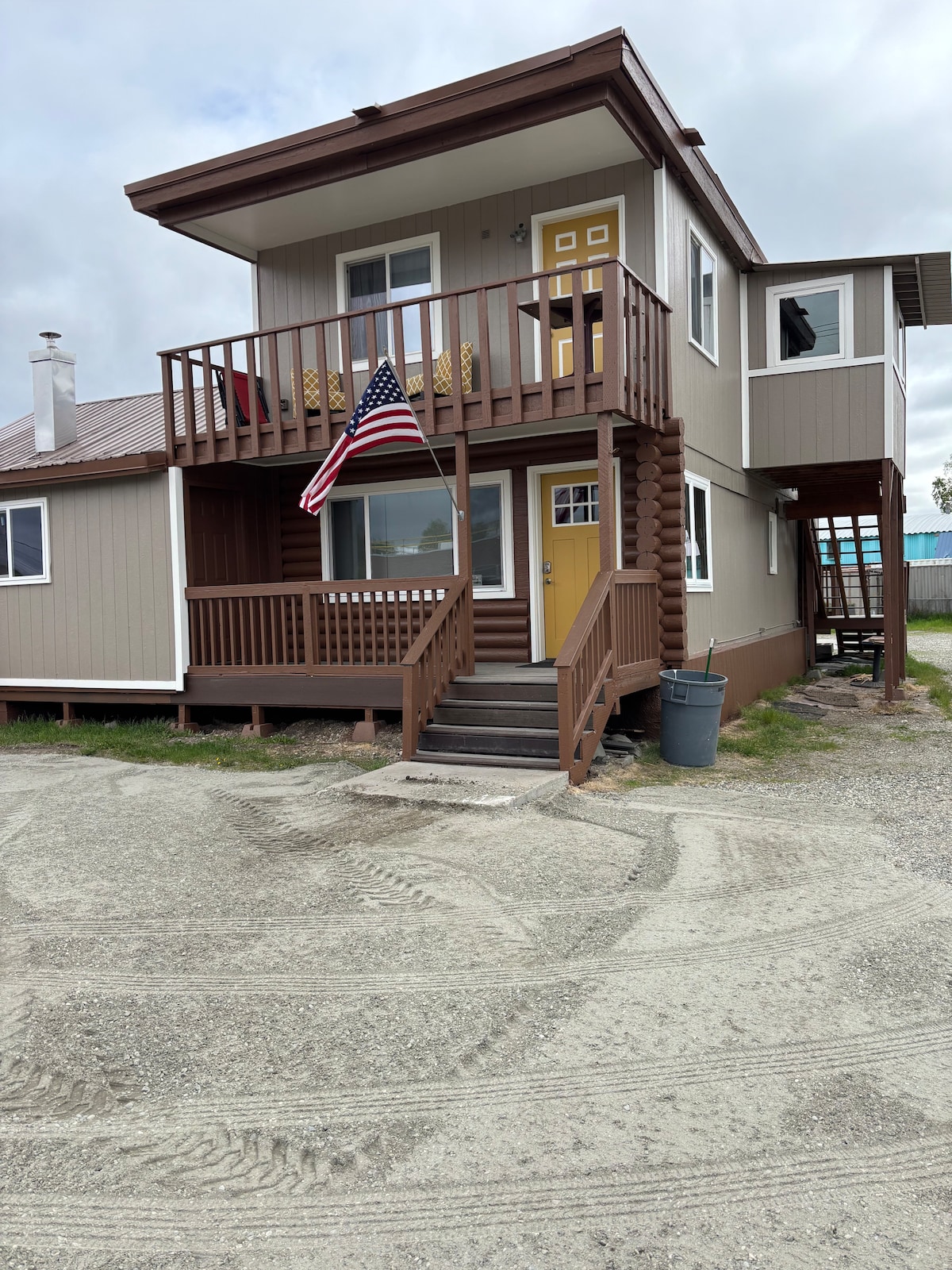 The exterior of a two-story apartment is showcased, featuring a welcoming entrance with a bright yellow door. A wooden deck is adorned with an American flag, and an ample gravel area provides parking. Light gray clouds hover above the structure, typical of Fairbanks' atmosphere.