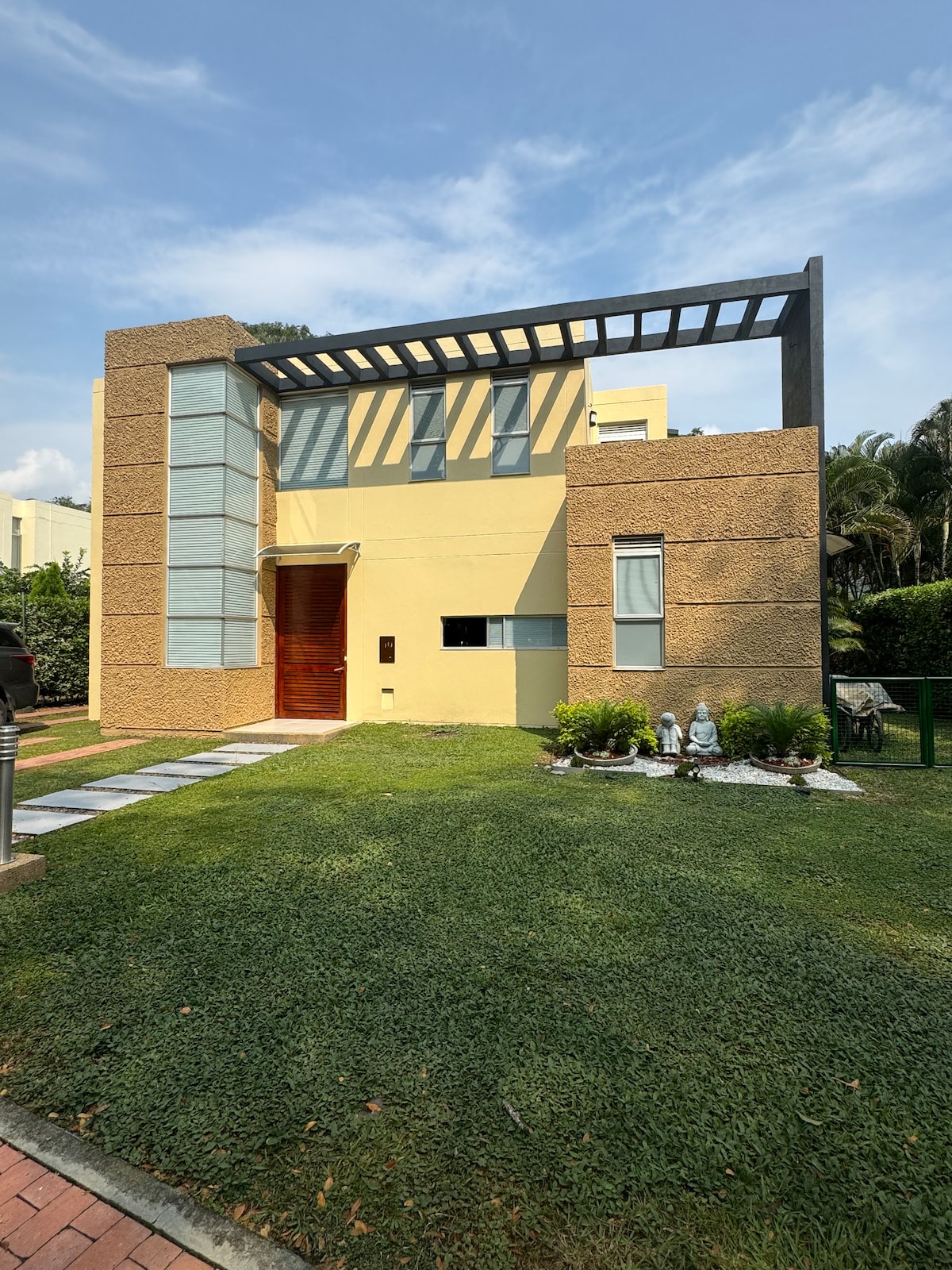 The exterior of the modern house features a combination of textured beige walls and large glass panels. A wooden front door is framed by a covered porch, leading to a well-maintained lawn with decorative plants. A stone pathway guides guests to the entrance.