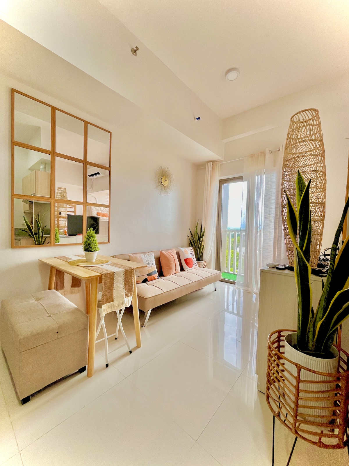 A bright living area features a modern design with a mirror reflecting natural light. A comfortable sofa and a small dining table are arranged with minimalistic decor. Potted plants add a touch of greenery, while a sliding glass door opens to a balcony, enhancing the space's openness.