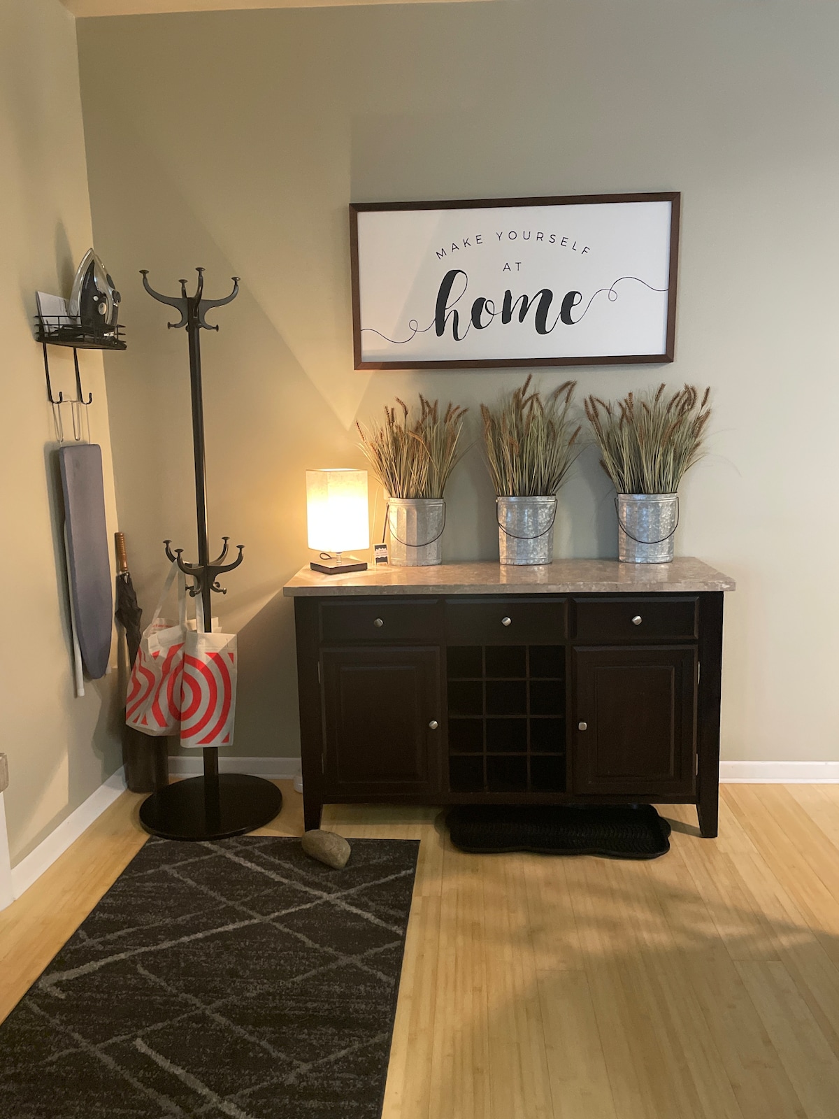 An entryway features a wooden sideboard with a marble top and a wine rack below. Metal vases with decorative grasses are placed on top, accompanied by a lamp providing soft illumination. A coat rack stands nearby, and a textured rug lays on the floor.
