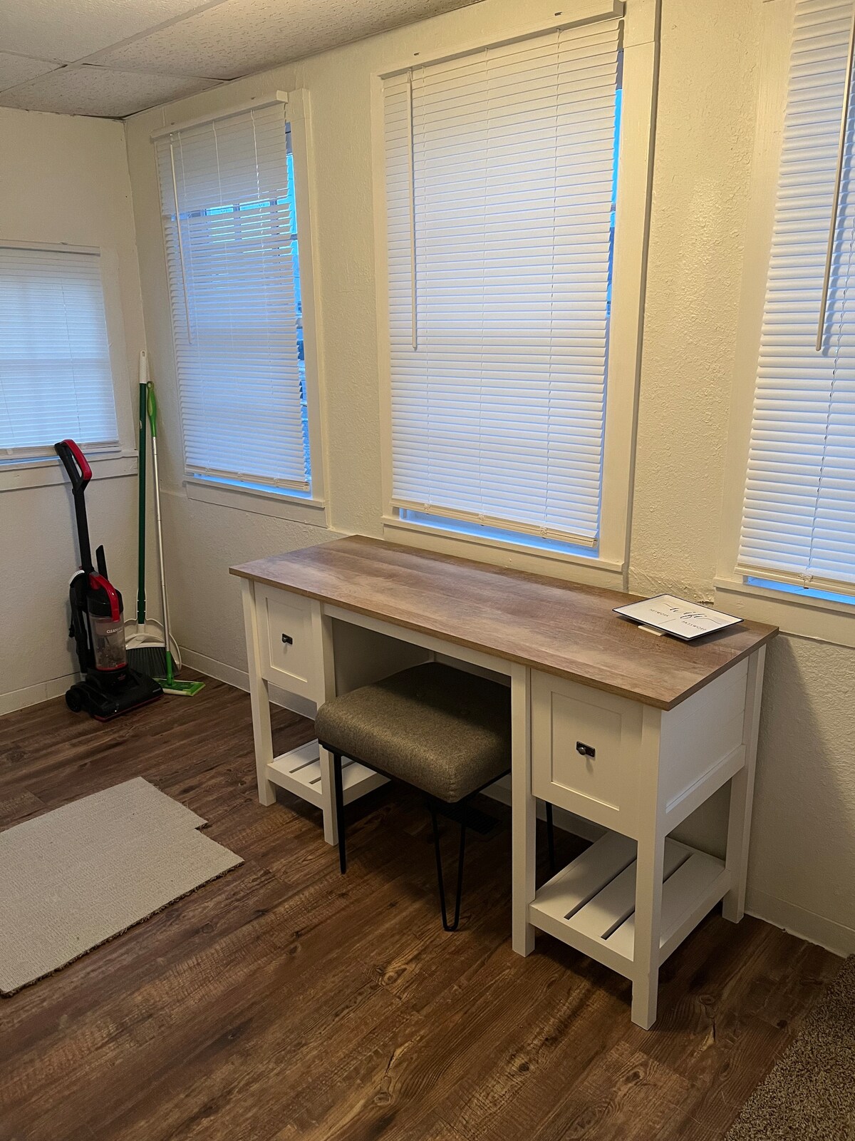 A workspace area is presented with a simple wooden desk and an upholstered chair. Natural light filters through two window blinds, creating a bright and open feel. A small vacuum cleaner and a soft rug are noted in the corner of the room.