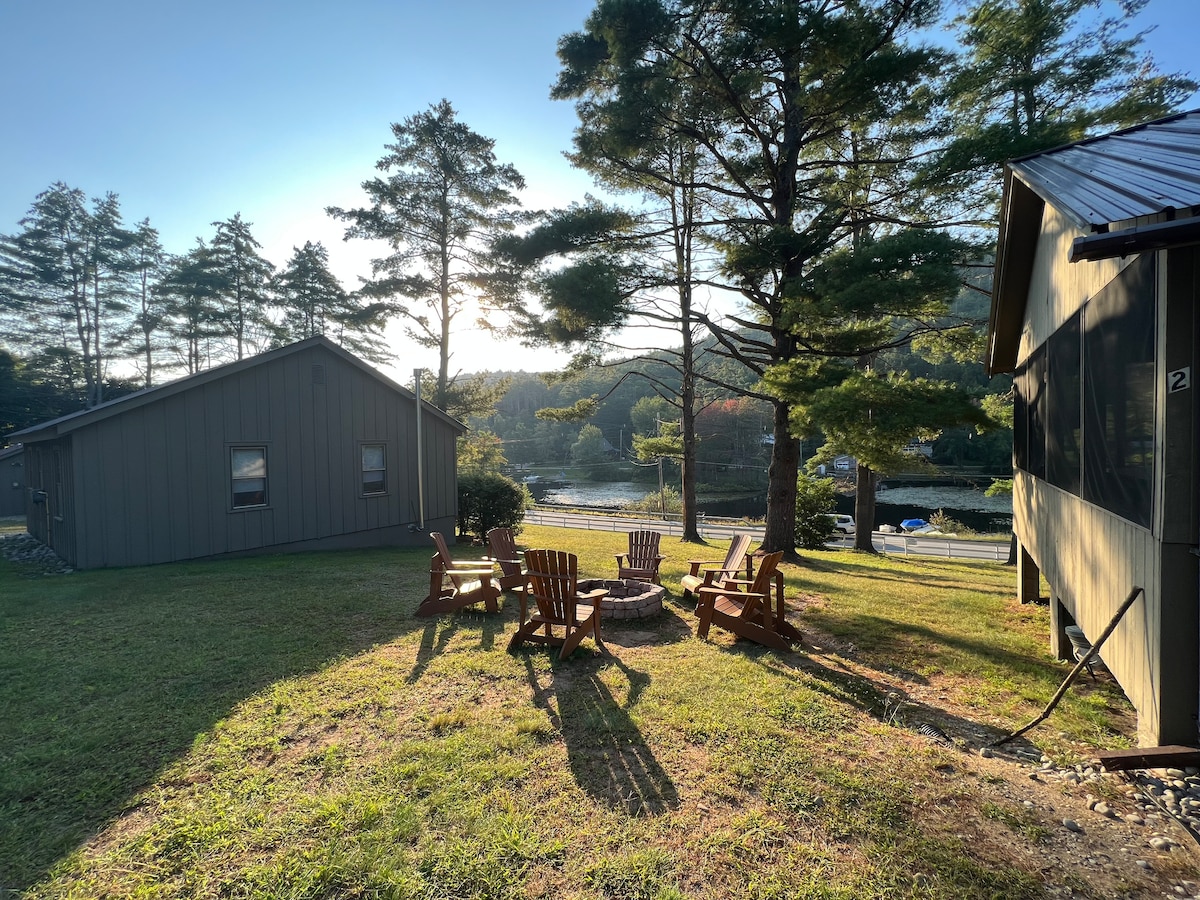 A serene outdoor space features Adirondack chairs arranged around a central fire pit on a grassy area. The backdrop includes trees and a glimpse of the creek, illuminated by soft morning light.