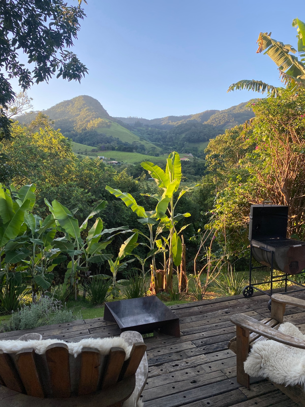 A wooden deck is surrounded by lush greenery, featuring tall banana plants and various trees. In the background, gentle mountains are visible under a clear blue sky. A barbecue grill is positioned on the deck, enhancing the outdoor cooking space.