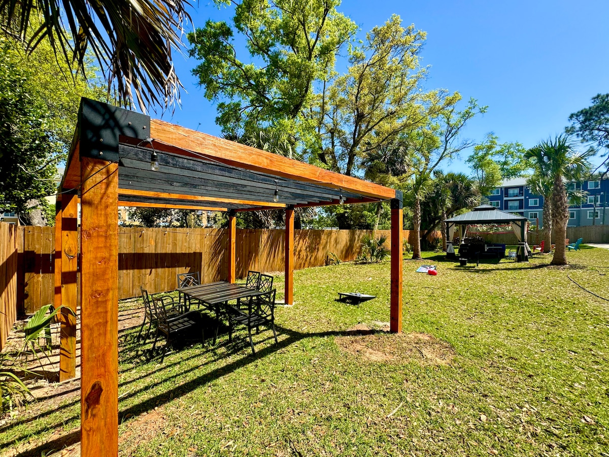 An outdoor patio scene showcases a wooden pergola providing shade over a dining table and chairs. A spacious lawn area is surrounded by a wooden fence, with palm trees and a gazebo visible in the background. The vibrant greenery suggests a serene and inviting atmosphere.