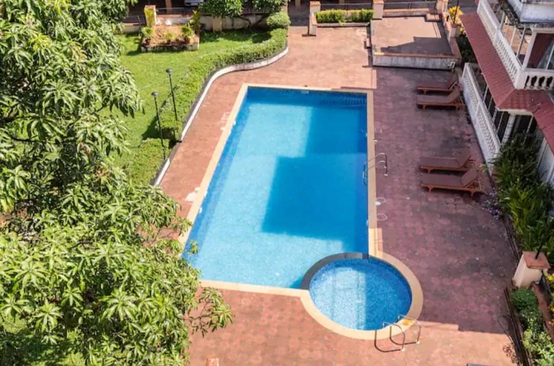 A large swimming pool is surrounded by a spacious patio area, featuring sun loungers. A smaller circular hot tub is situated adjacent to the pool. Lush greenery borders the area, creating a serene outdoor environment.