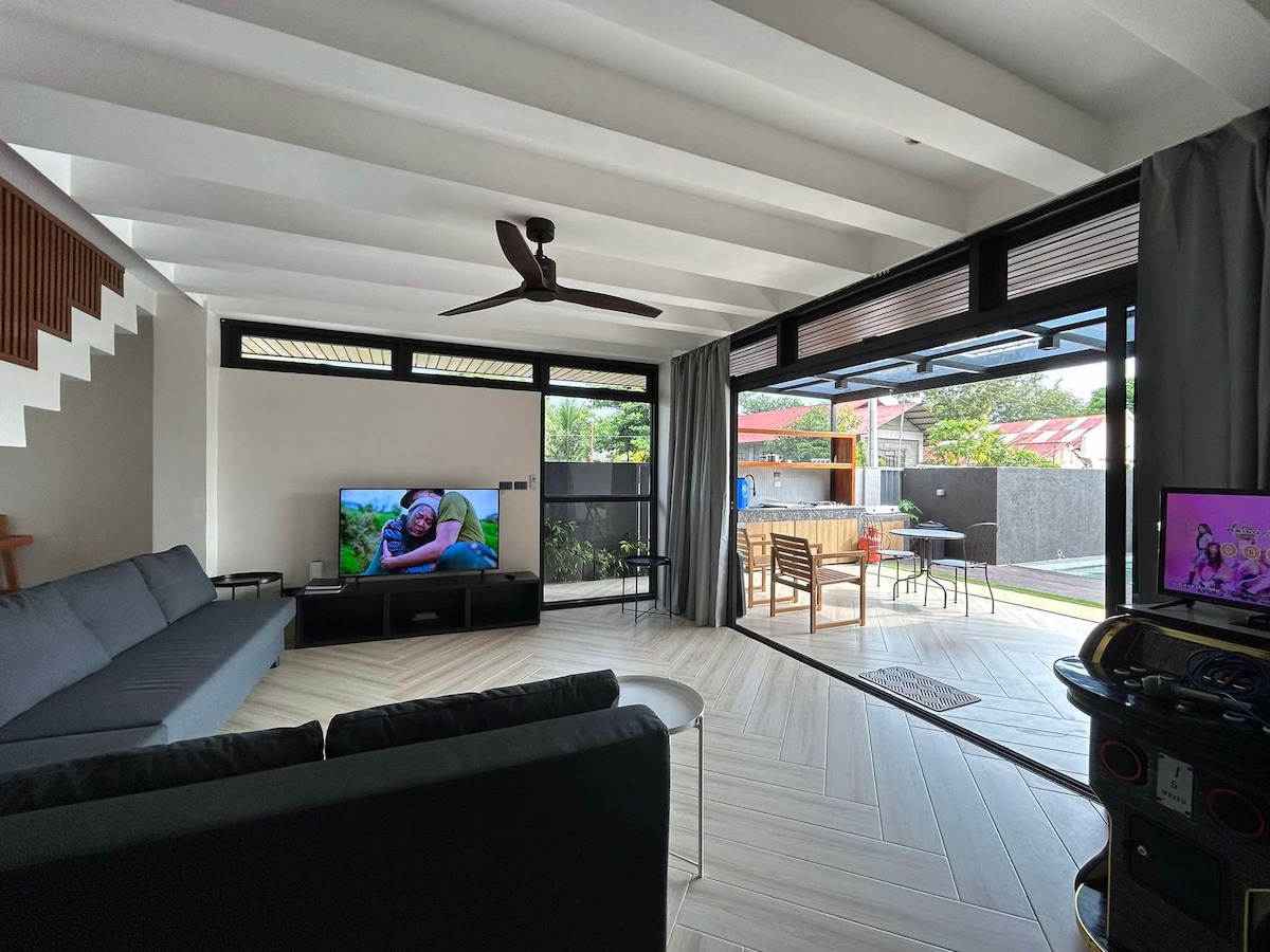 A bright living area features modern furnishings, including a dark sectional sofa and a flat-screen television. Large windows provide views of the outdoor space, while a ceiling fan enhances airflow. A cozy table is visible, along with outdoor seating through the open sliding doors.