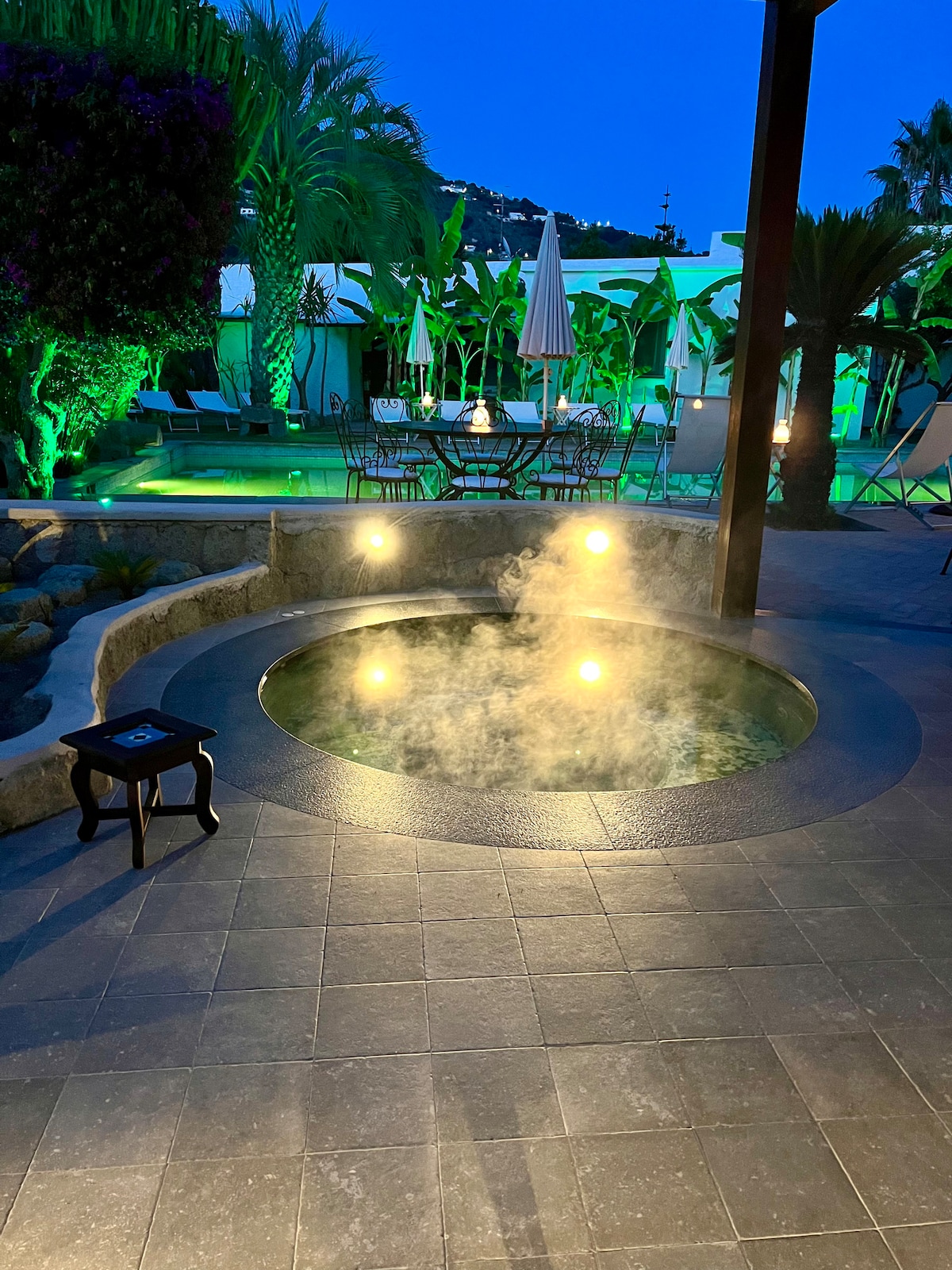 A warm, bubbling hot tub is surrounded by natural stone, steam rising gently into the evening air. Soft lighting highlights the lush greenery nearby, creating a serene ambiance. A small stool is positioned next to the tub, offering convenience for guests.